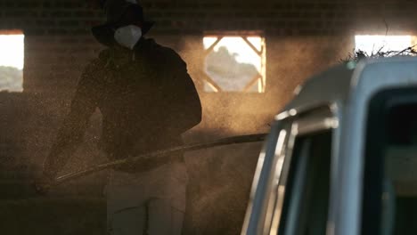 a man in a hat and wearing a dust mask loading hay onto the back on a farm vehicle with lots of dust in the air