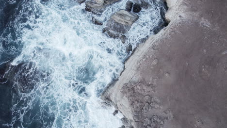 An-aerial-view-of-powerful-waves-crashing-against-the-rocky-coastline-of-Cyprus,-showcasing-the-raw-energy-of-nature