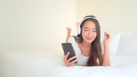 Barefoot-young-pretty-girl-lying-on-bed-in-bedroom-swaying-while-listening-music-using-headphones-and-smartphone