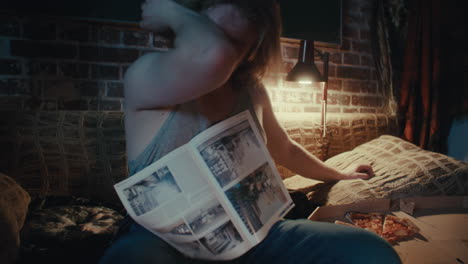 man reading newspaper and eating pizza in a bedroom