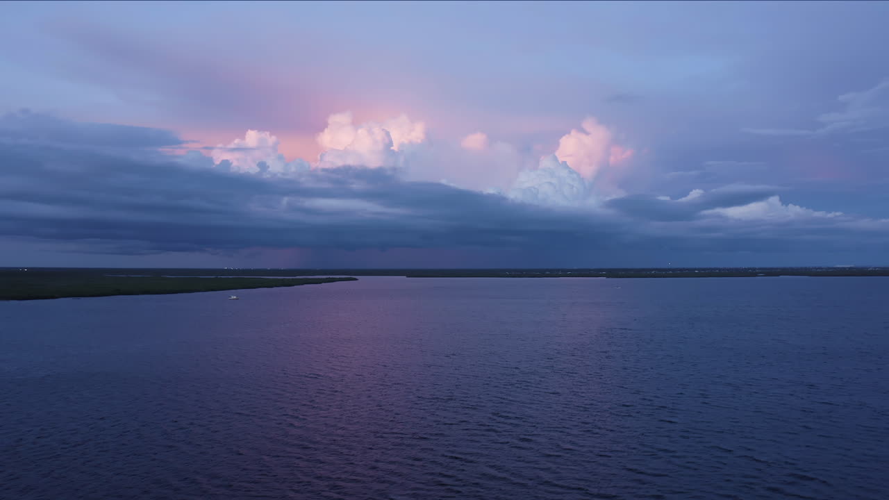 La delicata luce serale accarezza un lago tranquillo mentre imponenti nuvole temporalesche si illuminano di rosa al sole calante, creando un contrasto pacifico tra l'acqua calma e il tempo che si prepara.