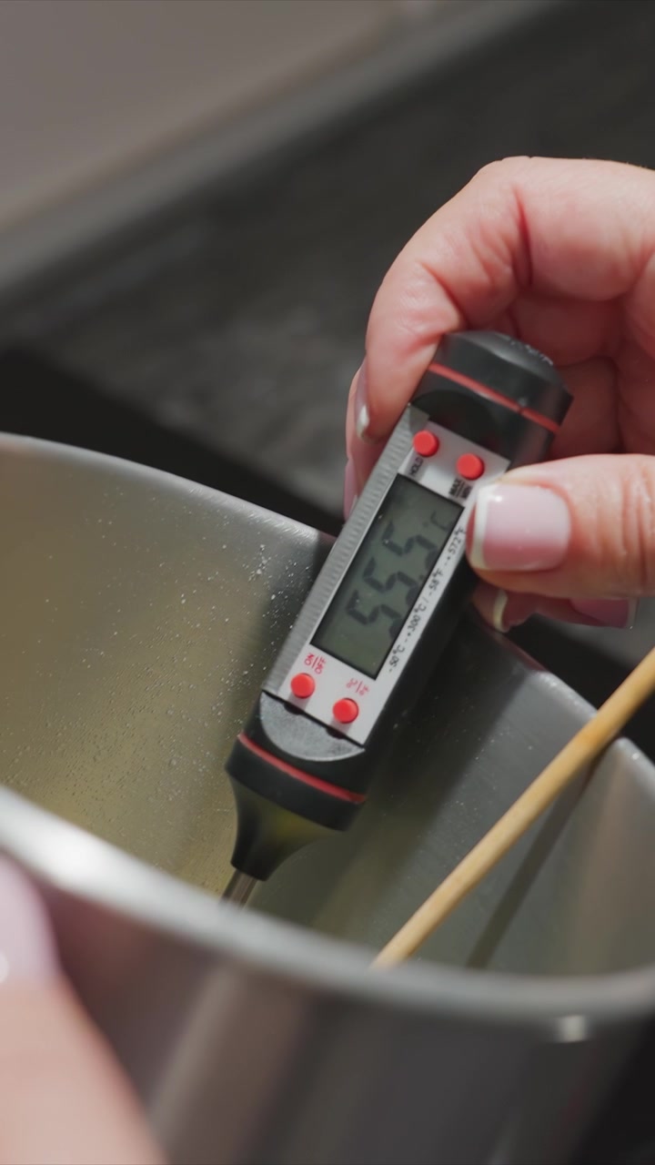 Primo piano di una mano che tiene un termometro digitale in un liquido bollente per misurare la temperatura. La scena mostra le dita della persona con unghie curate che afferrano il dispositivo mentre visualizza letture che cambiano.