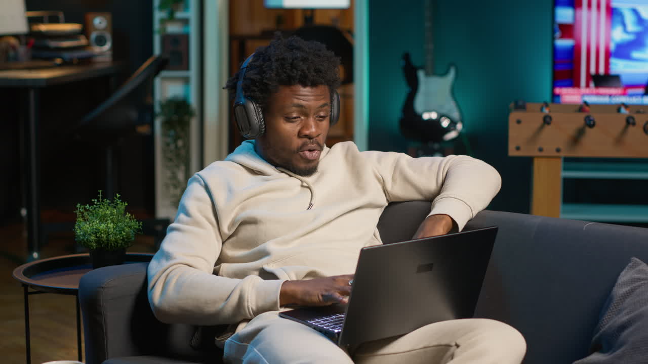 Freelancer listening to a podcast and being focused on online work, putting on headphones while seated on the couch in his living room. Productivity in a casual and flexible home. Camera A.