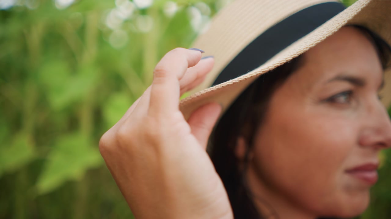 Primo piano di una donna che si sistema il cappello di paglia con una mano all'aperto. L'attenzione rimane sull'interazione tra la donna e il suo accessorio, evidenziando texture naturali come il materiale intrecciato.