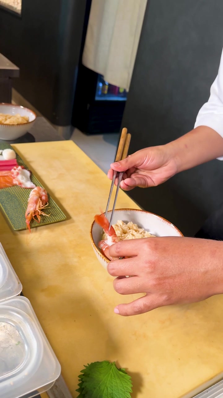 Koki profesional menata udang yang sudah dimasak di atas mangkuk nasi dengan sumpit di konter sushi kayu.