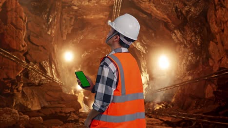 back view of asian male engineer with safety helmet working on a green screen smartphone and looking around while standing in underground mine tunnel