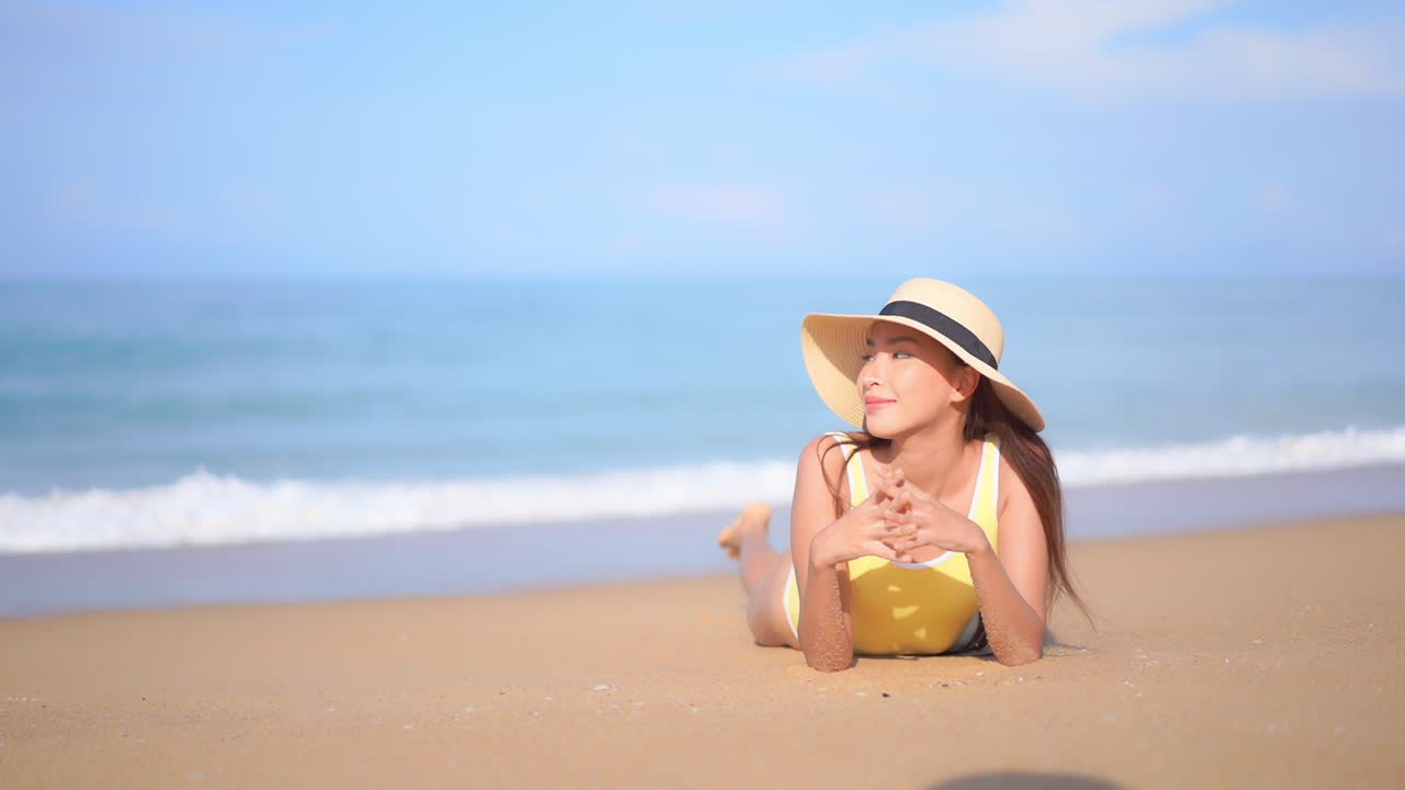 거품 파도가 모래 위로 굴러갈 때 바다 근처의 모래 해변에서 팔꿈치에 기대어 누워 쉬고 있는 노란색 수영복과 햇빛 모자