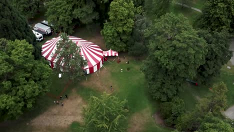 people walking in the park around the circus tent surrounded by the lush green trees in summer