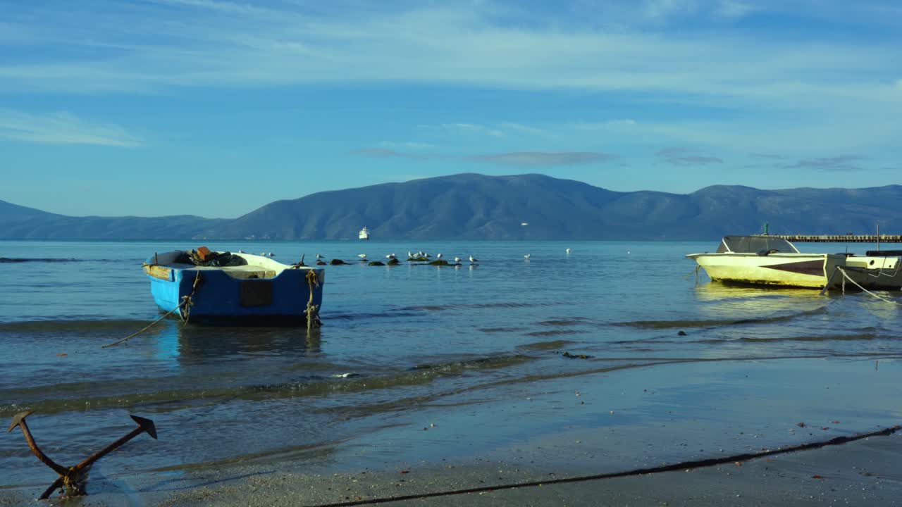 barcos de pesca en la orilla del mar con ancla hundida en la arena, gaviotas y fondo de montaña rocosa bajo un cielo nublado