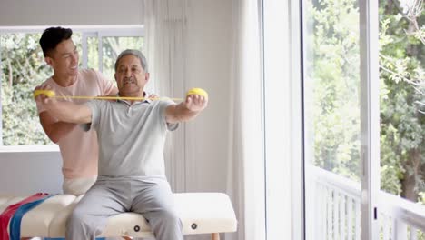 Diverso-Fisioterapeuta-Masculino-Asesorando-Y-Paciente-Masculino-Mayor-Usando-Banda-De-Ejercicio,-En-Cámara-Lenta