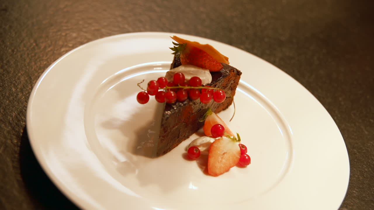Slice of fancy chocolate cake garnished with strawberries, red currants, whipped cream, and a caramel crisp presented elegantly on a white plate. Push in close up.