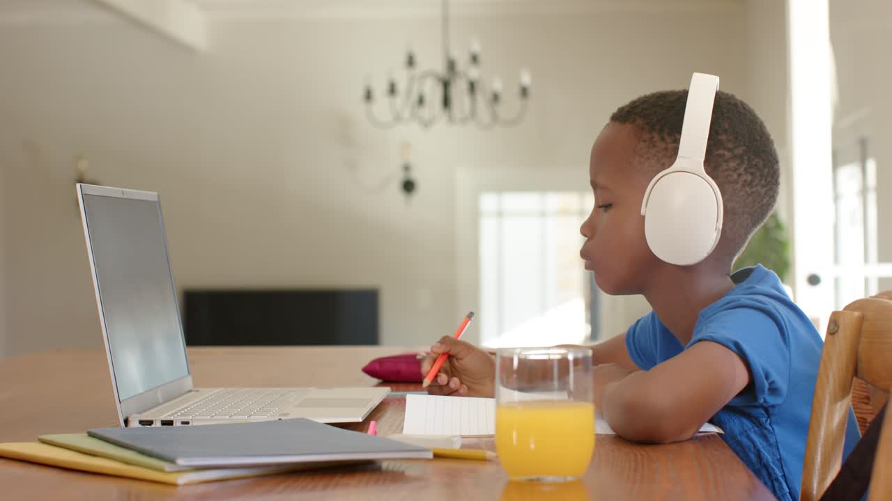 African American boy in headphones responding on laptop, writing pencil at dining table, copy space