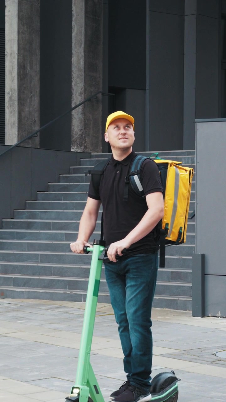 A delivery man rides an electric scooter down a city street with a yellow delivery bag on his back