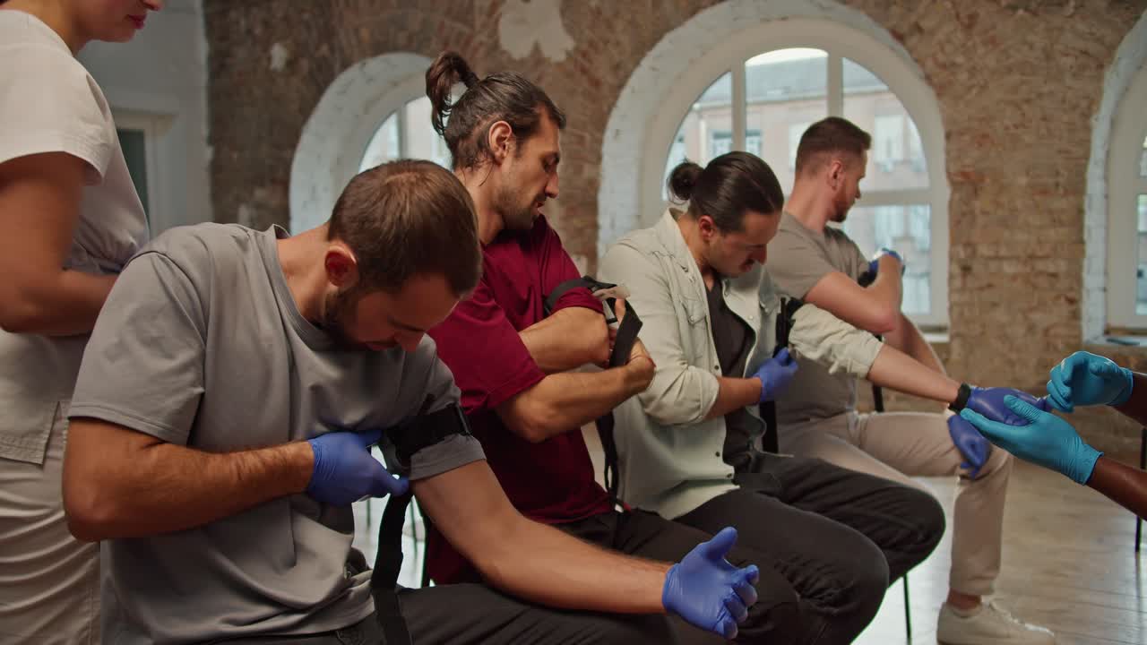 Close-up shooting: a group of people learn to work with a medical tourniquet under the guidance of a dark-skinned doctor in a blue medical uniform during a practical medicine class, a professional nurse girl in a white medical uniform checks the correct application of tourniquets