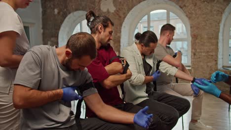 Close-up-shooting:-a-group-of-people-learn-to-work-with-a-medical-tourniquet-under-the-guidance-of-a-dark-skinned-doctor-in-a-blue-medical-uniform-during-a-practical-medicine-class,-a-professional-nurse-girl-in-a-white-medical-uniform-checks-the-correct-application-of-tourniquets