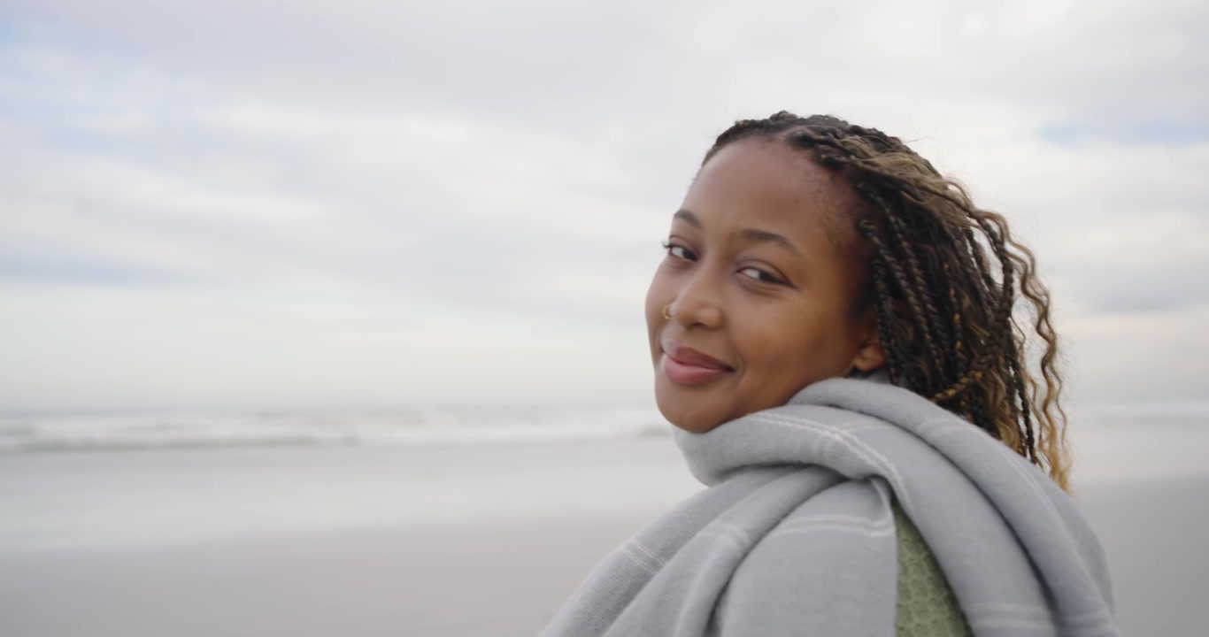 Adult African woman breathing breeze on shore, relaxing, pulling blanket over sweater, copy space