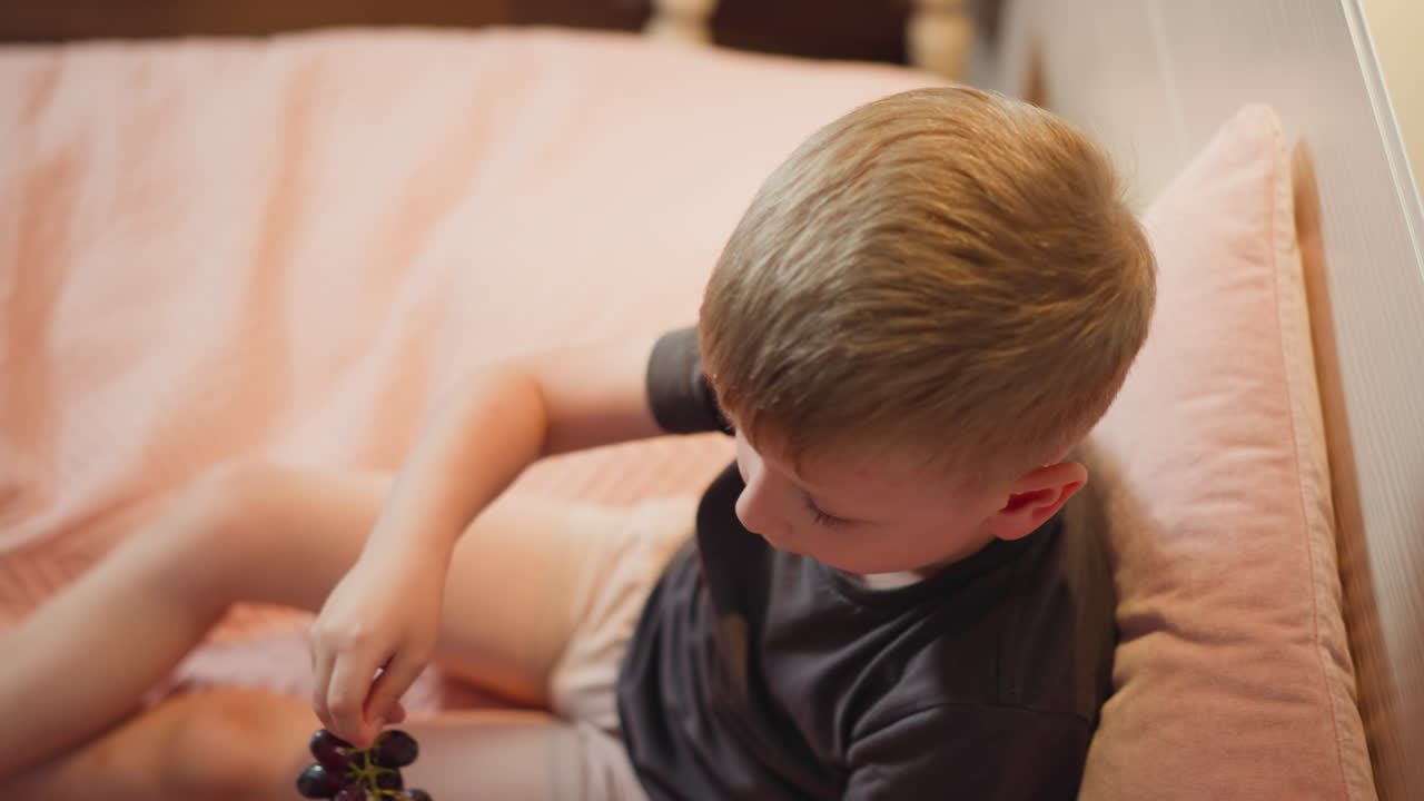 Top view of young boy sitting on pink bed holding grape bunch while eating slowly with focused expression, soft lighting captures peaceful indoor moment of healthy snacking