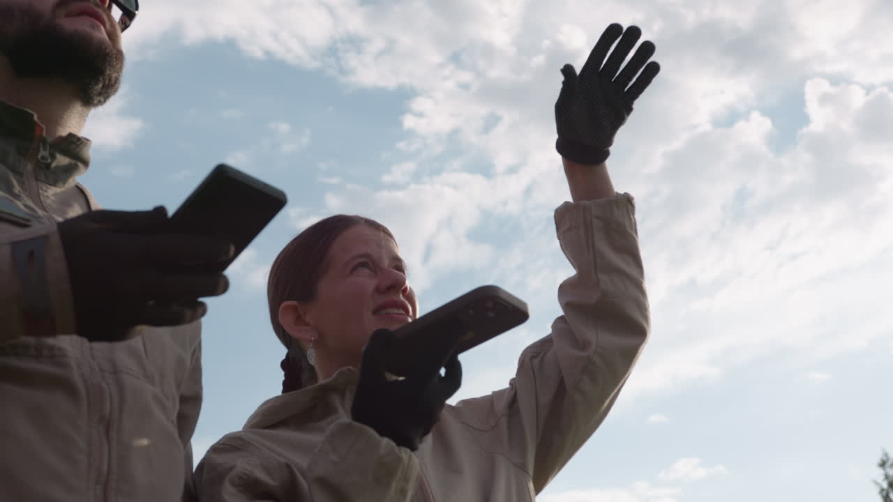 close up of man and woman holding phone and observing sky, gloved hands gripping device while gaze follows clouds, sun flare catching cloud edges, focus and anticipation over forest backdrop