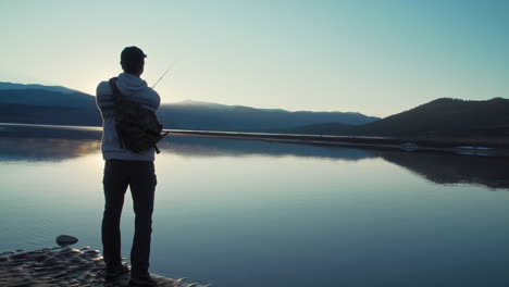 Medium silhouette shot of a fisherman casting his lure into a still, glassy, lake in front of gorgeous, snow-capped mountains