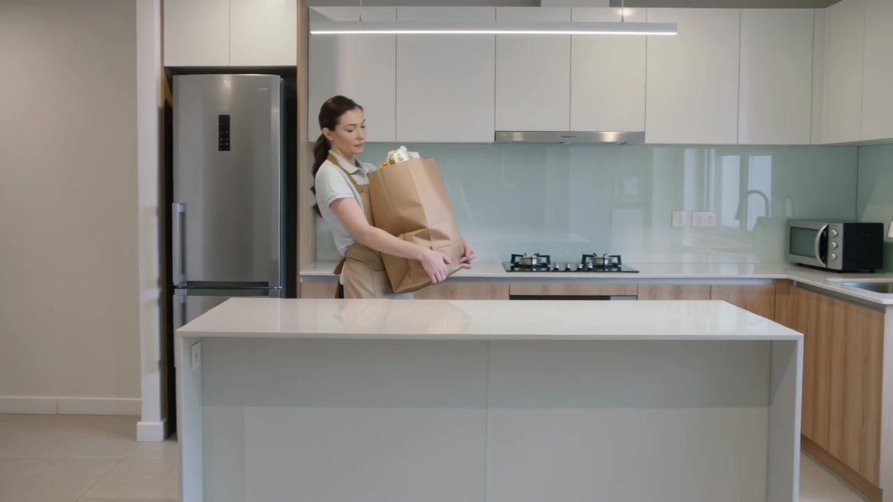 Una donna entra in cucina con un grembiule color cachi, tenendo una borsa di carta, la posiziona sull'isola e controlla la spesa