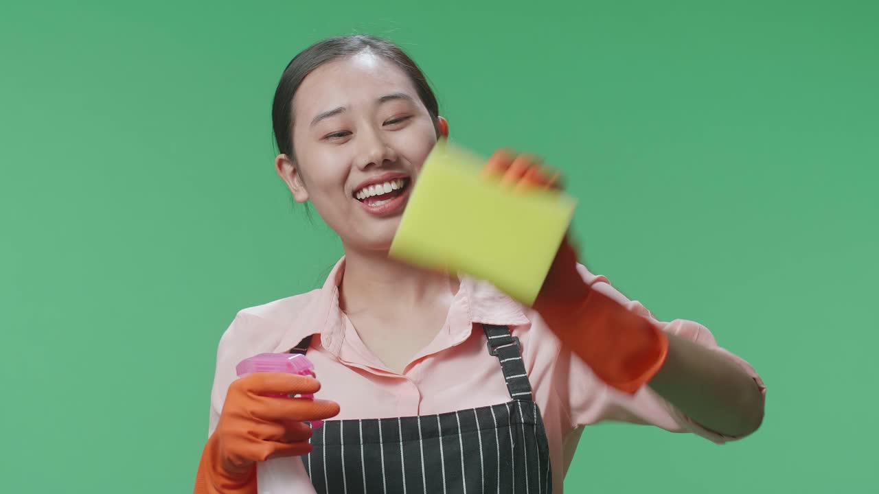 초록색 스크린 스튜디오에서 스프레이와 부 스폰지로 유리잔을 는 아프론을 입은 미소 짓는 아시아 여성 집주인