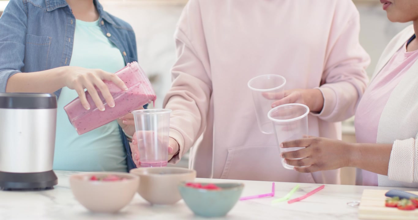キッチンで、ブレンダーのピッチャーを持ち上げ、ピンクの飲み物をカップに注ぎ分けている多様な女性たち。