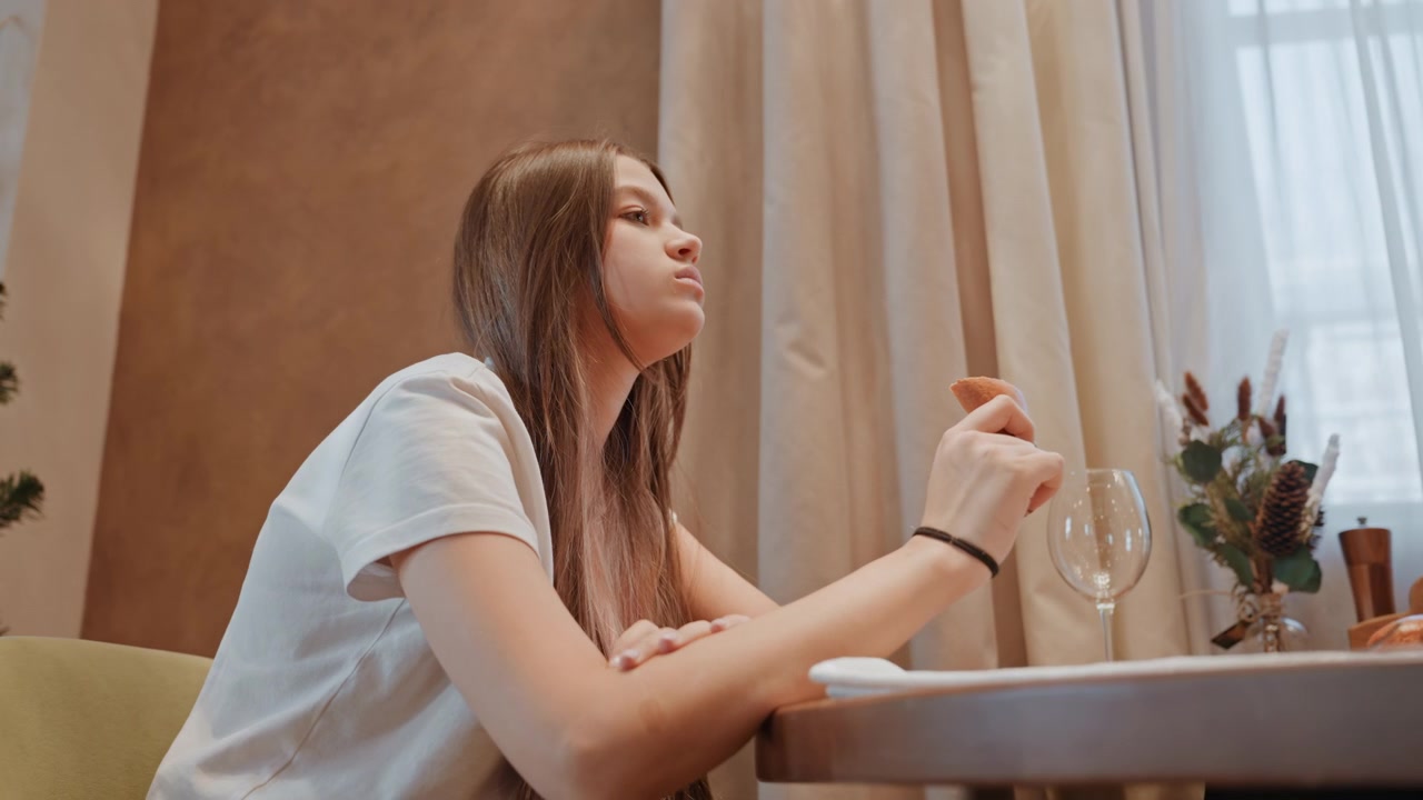 Young black woman sitting at table by window, braided hair holding macaron, contemplative expression, soft sunlight through sheer curtain, warm neutral decor, small pine plant and wine glass on table