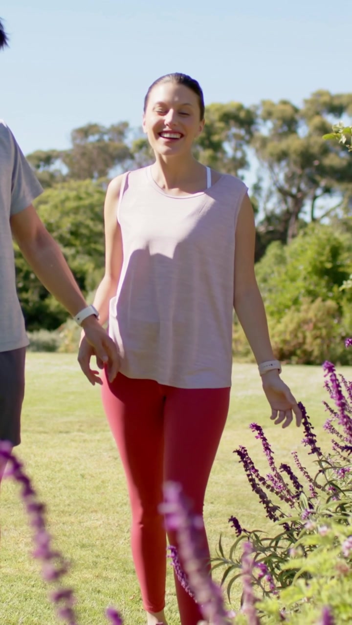 Video vertical: Pareja caminando de la mano pasando lavanda en un parque, mujer con top rosa usando reloj.