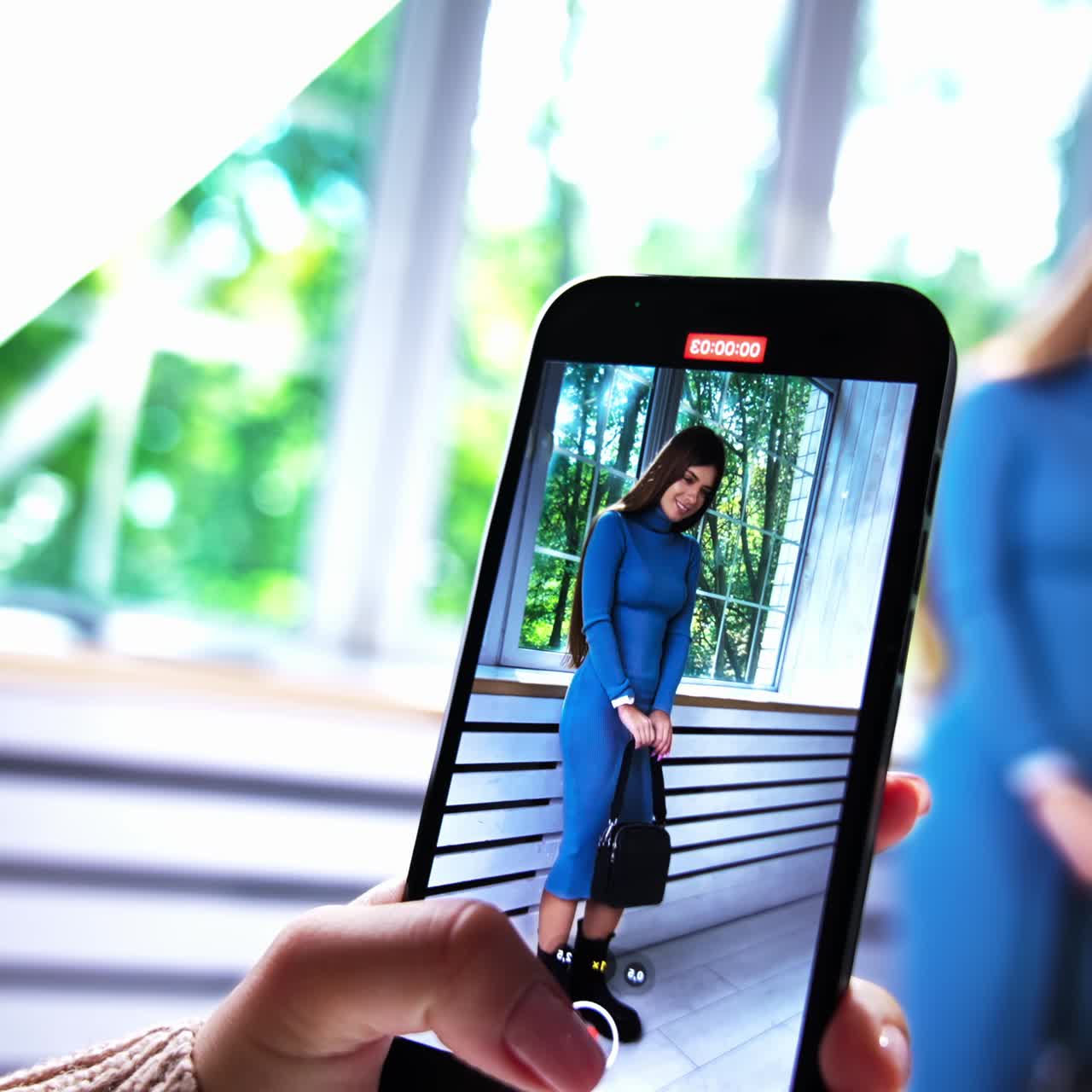 Taking video of a model posing on a smartphone. Brunette woman in blue dress, black boots and with black bag displaying fashionable look