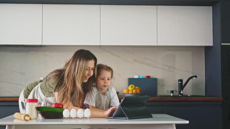 pretty-mom-stands-in-kitchen,-leaning-over-her-daughter