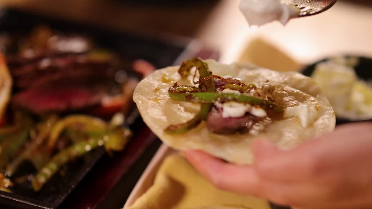 Hands prepare a steak fajita with grilled vegetables and sour cream on a tortilla, under warm lighting, with shallow depth of field and close-up perspective