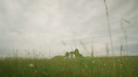 in a field of tall grasses, a painter wearing a hat and checkered shirt is intensely focused on creating art on a board under a cloudy sky. nearby, a woman sits comfortably on a chair