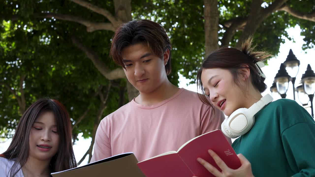 Three Asian students smile and talk while reviewing notebooks under trees in natural daylight