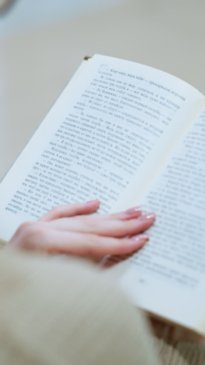 vista trasera de cerca de una dama con uñas pulidas volviendo a la siguiente página del libro, borrón parcial de los vasos visibles en un entorno bien iluminado, enfoque suave en el texto y manos delicadas
