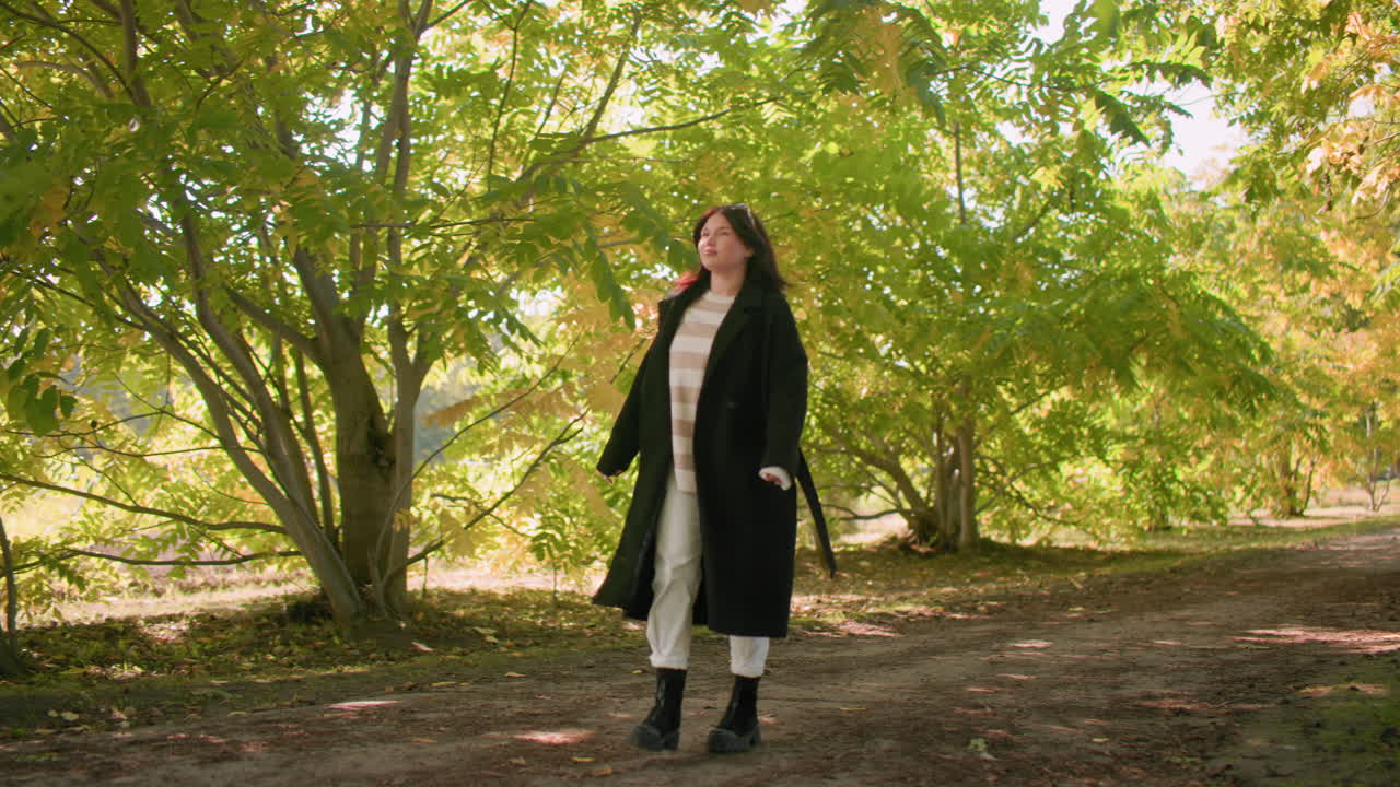 Donna elegante che cammina con maestosità lungo il sentiero di un arboreto, cappotto invernale e stivali neri, sguardi pensosi verso la volta frondosa, luce calda autunnale e ombra soffusa, passo rilassato, atmosfera tranquilla all'aperto