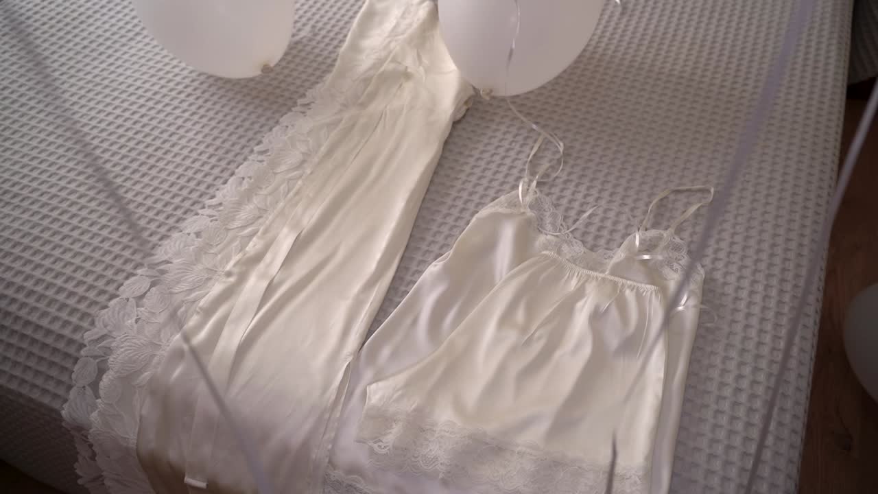 Un abito bianco e una camicia da notte sono appoggiati ordinatamente su un letto. Nelle vicinanze, dei palloncini fluttuano, aggiungendo un tocco festoso alla stanza. Una luce soffusa illumina lo spazio.