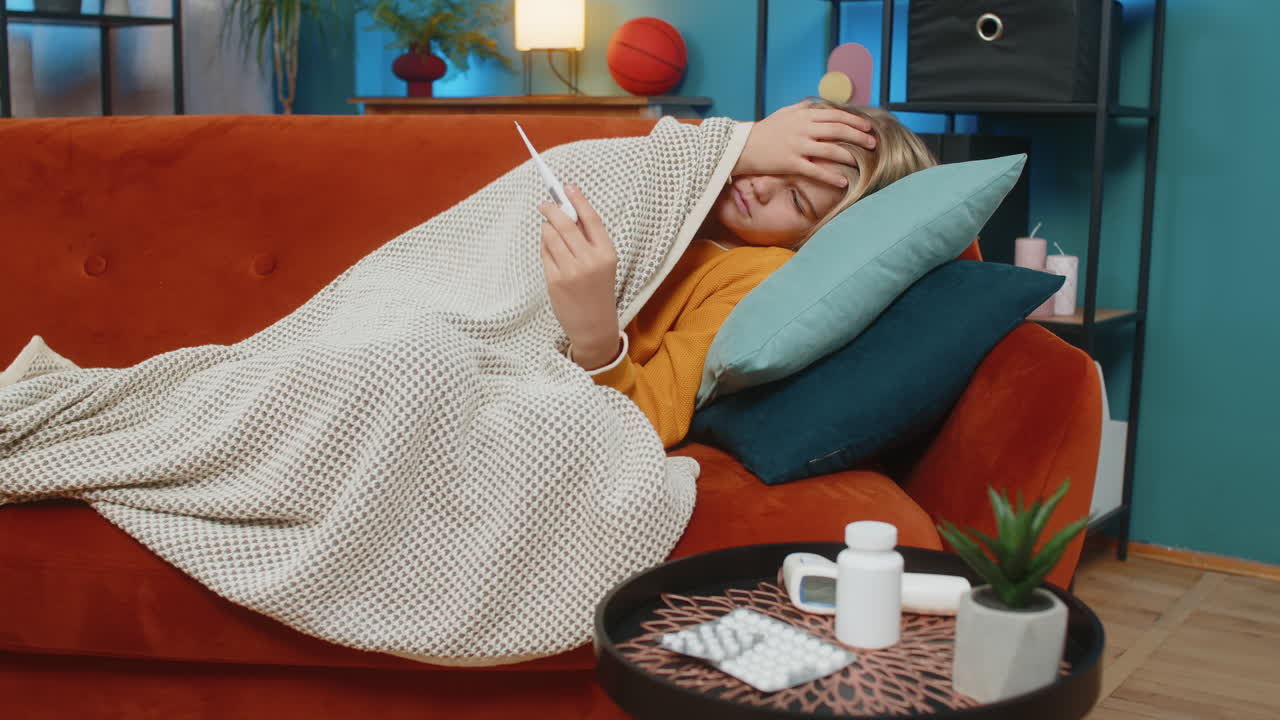 Teenager boy sick with fever lying on sofa checking high temperature on thermometer sad tired home