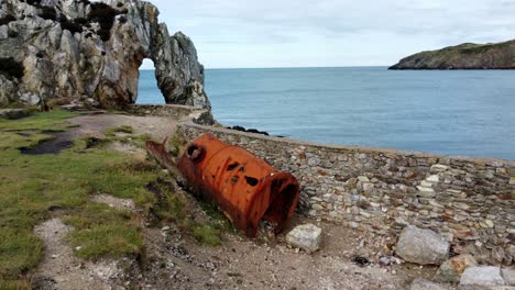 vista aérea que rodea el tanque industrial oxidado rojo para revelar el arco de roca en la costa galesa