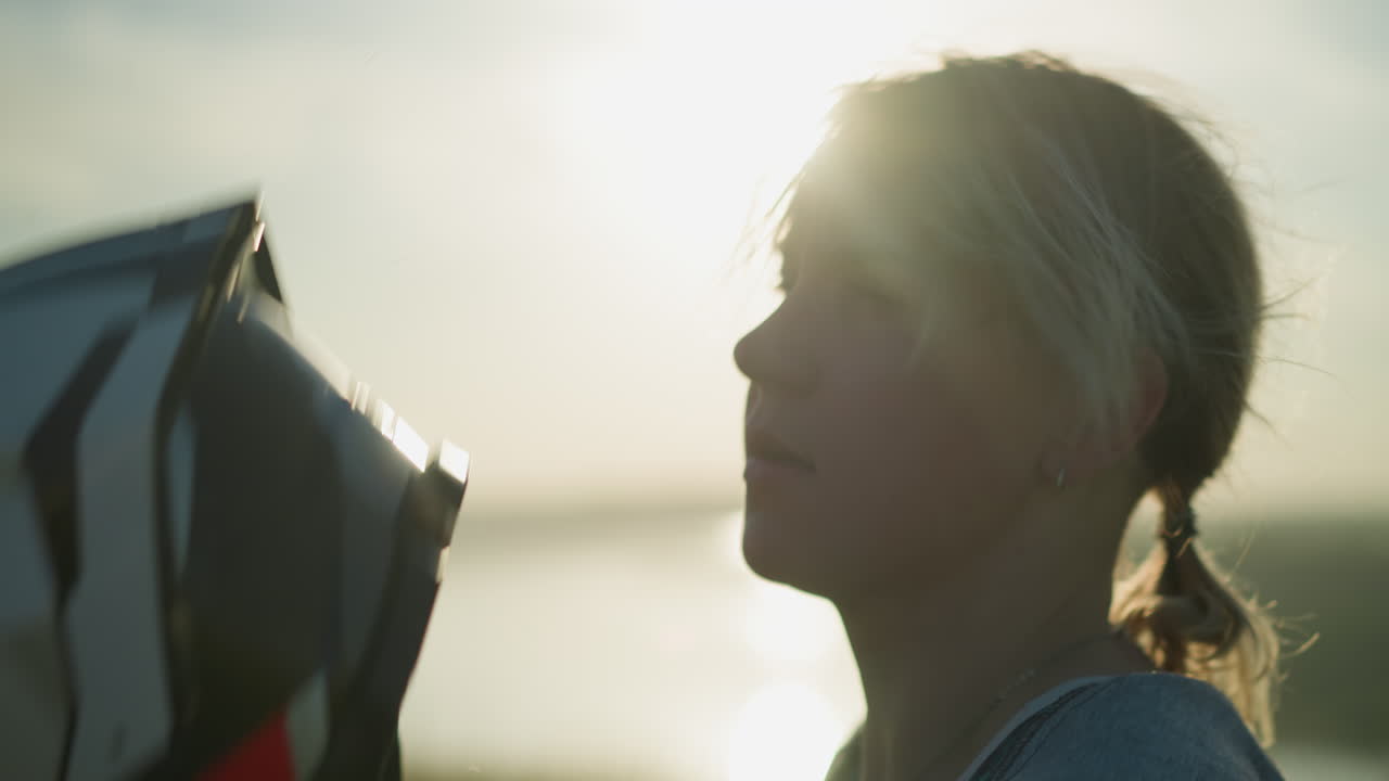 молодая женщина начинает снимать шлем одной рукой, а другой осторожно стирает волосы назад, в то время как солнечный свет отражается на ней