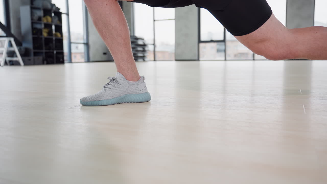 Male trainer leg view stretching both legs in side lunge on wooden gym floor with sneakers showcasing muscle tone under large windows and bright studio lighting in modern fitness environment