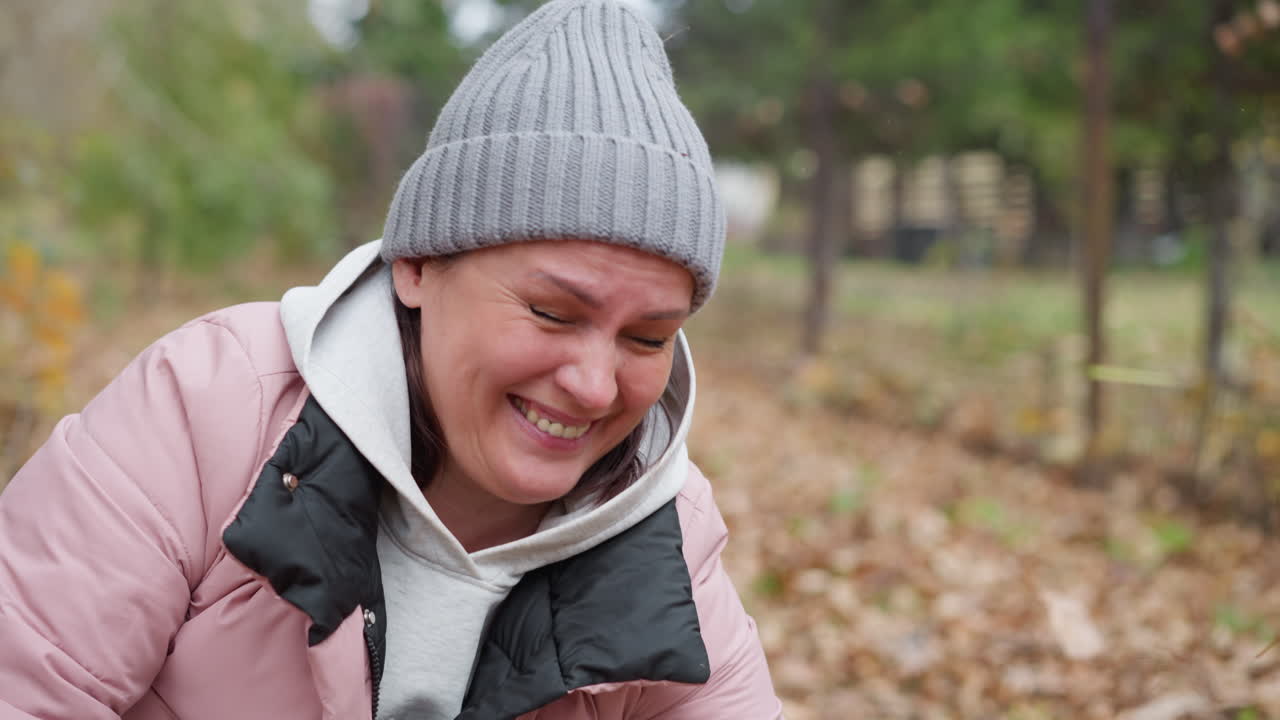 mujer sonriente con chaqueta rosa y gorra gris se inclina al aire libre mientras el follaje seco se vierte juguetónamente sobre ella, rodeada de vibrantes colores de otoño y paisajes naturales