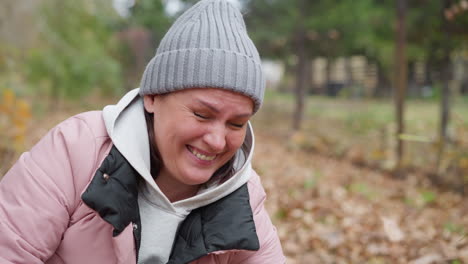 smiling woman in pink jacket and grey beanie bends down outdoors as dry foliage is playfully poured on her, surrounded by vibrant autumn colors and natural scenery