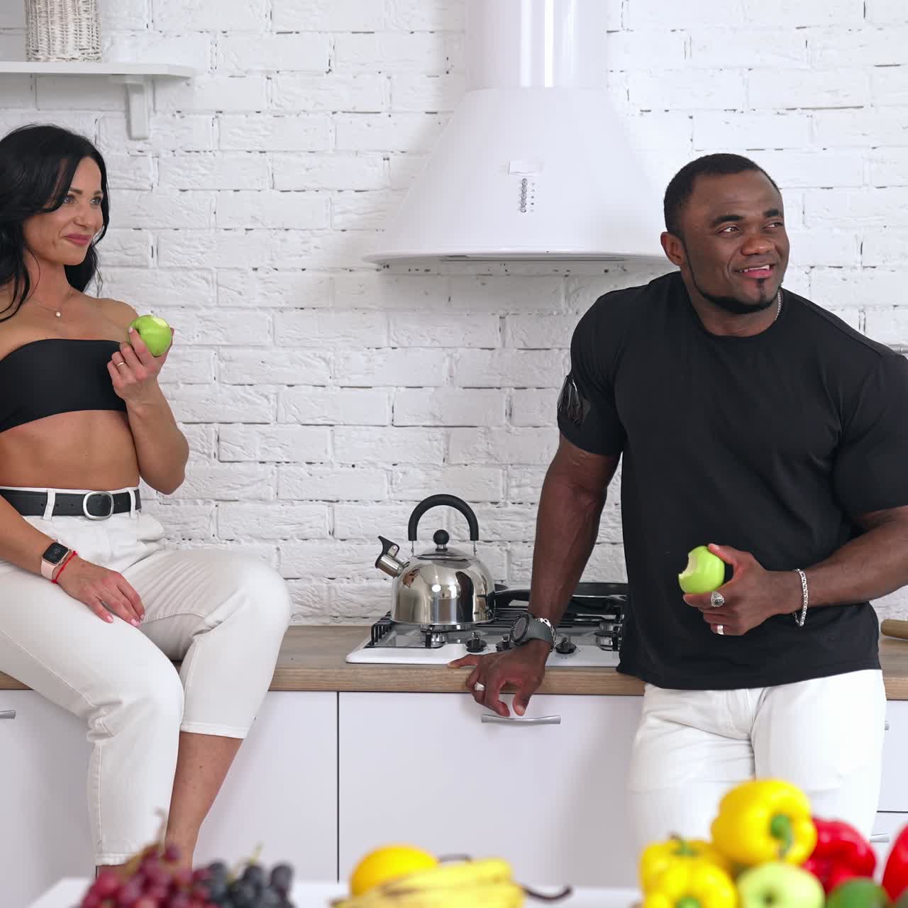 Interracial couple eating green apples in the kitchen. Woman sitting on the table and man standing nearby. Both look at the window and smile