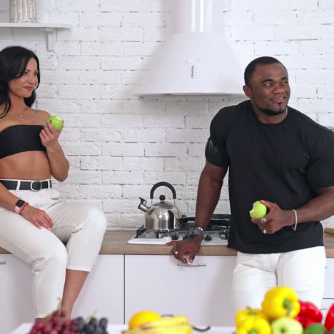 interracial couple eating green apples in the kitchen. woman sitting on the table and man standing nearby. both look at the window and smile