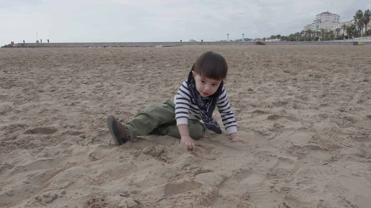Un bambino gattona e poi si siede sulla sabbia mentre la madre si avvicina per togliergli la sciarpa.