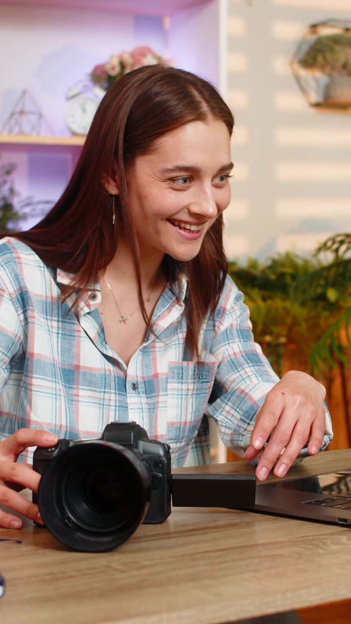 Giovane donna al tavolo di casa che modifica contenuti fotografici e video sul portatile con la fotocamera per lavoro creativo da casa.