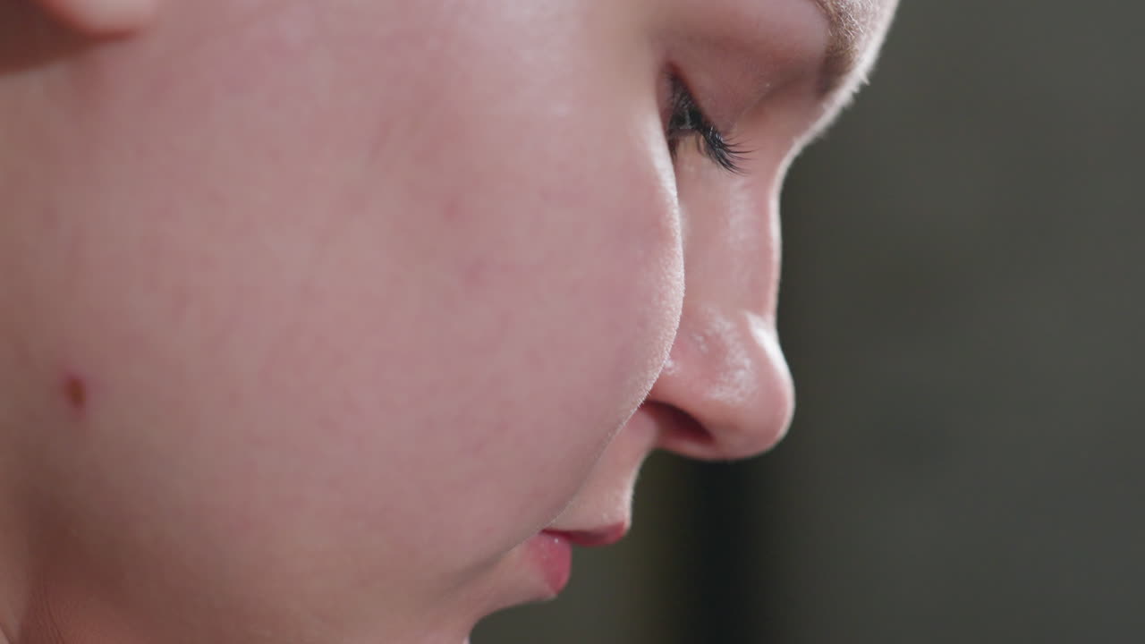 Side close view of girl indoors, spotless skin under soft light, eyes lowered with calm focus, serene profile implying careful task out of frame, lashes with smooth complexion