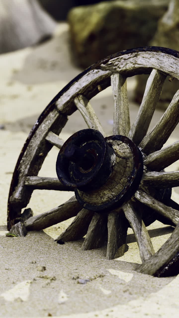 Old wooden wheel partially buried in sandy beach surrounded by rocks