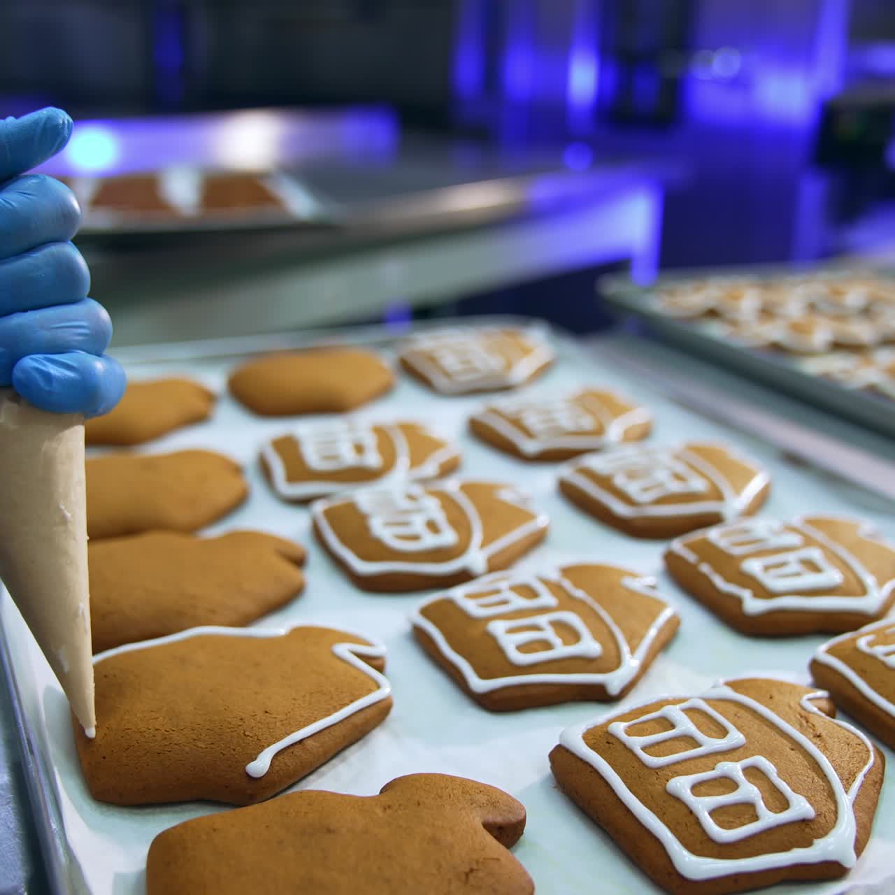 Confectionery worker holding a pastry bag and squeezing white cream on the cookies. Gingerbread houses close up. Trays with pastry in blur at background