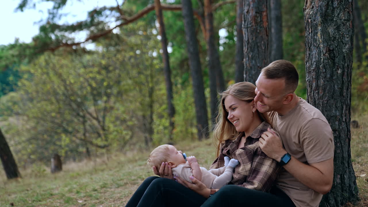 Man embraces his wife holding a baby boy. Caucasian couple sits near the tree smiling to their child with pacifier.